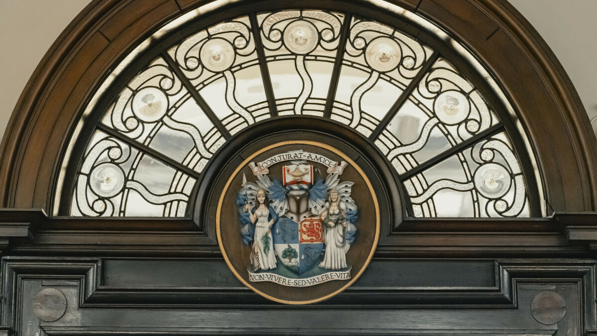 Semi circle of glasswork above doorway in College Hall. It curves around wood carving of College crest